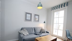 Een woonkamer met een grijze bank, twee ingekaderde foto’s aan de muur, een houten salontafel, een vloerlamp en een raam met een gestreept gordijn.