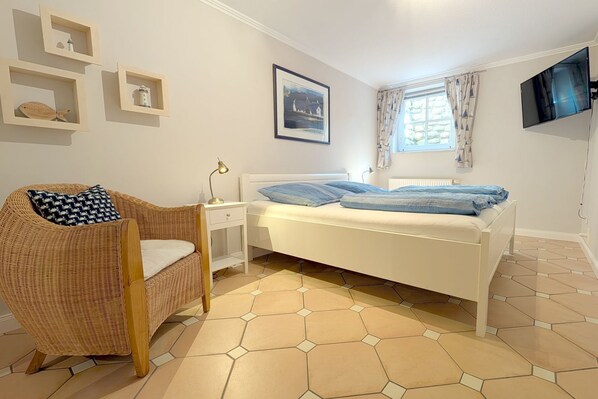 A bedroom with a bed, a wicker chair, a bedside table, a lamp, a framed picture, a window with curtains, and a TV mounted on the wall.