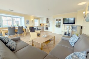 A modern living room with a sofa, dining table, chairs, and a kitchen area with a TV and speakers.