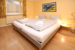 A double bed with white linens and towels, a bedside table with a lamp, a window with curtains, and a painting on the wall.