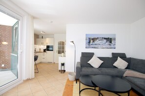 A modern living area with a grey sofa, a small round table, and a kitchen in the background. - Osterstraße 1, Whg. 2, Haus Flora - Apartment in Wyk/ Südstrand with approx. 60m² (Wyk auf Föhr)