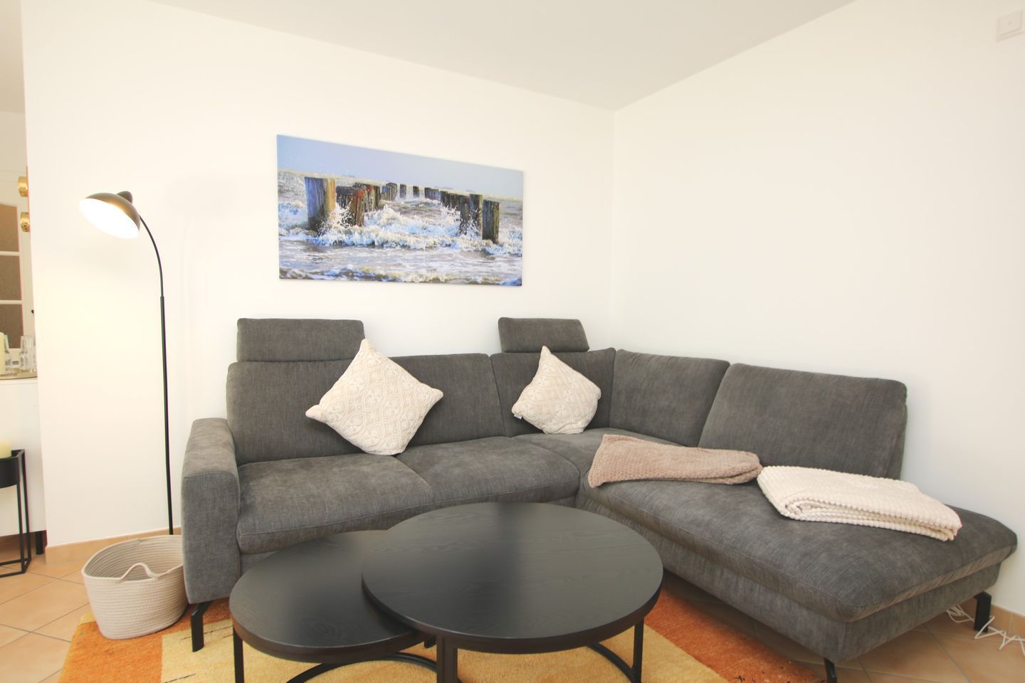 Un salon moderne avec un canapé d’angle gris, deux tables basses noires et un tableau représentant un paysage enneigé.