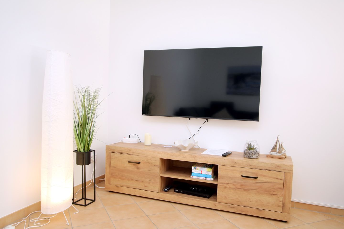 Una sala de estar moderna con un televisor de pantalla plana montado en la pared, un mueble bajo para televisión de madera con un control remoto, una planta pequeña y un objeto decorativo.