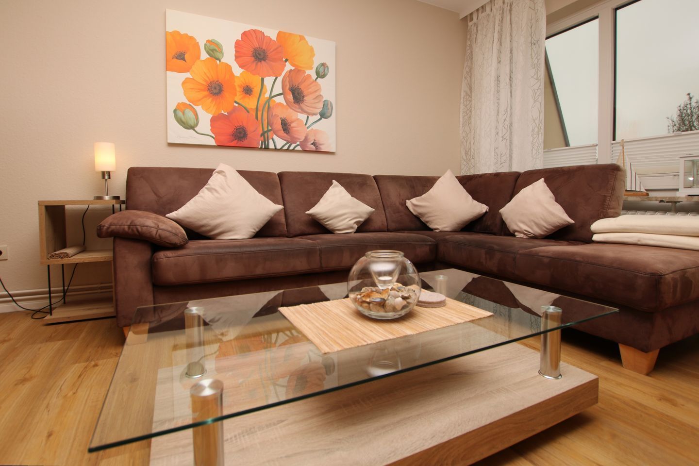 Un canapé en cuir marron avec des coussins blancs, une table basse en verre sur laquelle repose un bol décoratif, et un tableau représentant des fleurs oranges accroché au mur.