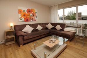 Uma sala de estar moderna com um sofá em tons de castanho, uma mesa de centro de vidro, uma pintura de flores e vista através de grandes janelas.