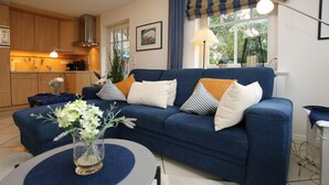 A living room with a sofa, a table with a vase, a kitchen in the background, and a window with curtains.
