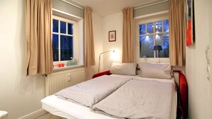 A double bed with a red headboard, beige curtains, a bedside lamp, and a window with a view of trees.