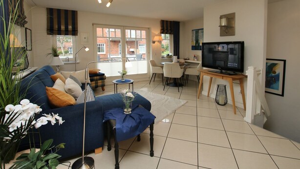 A living room with a sofa, coffee table, and TV. There is a dining area with chairs and a table. The room has large windows and a view of buildings outside.