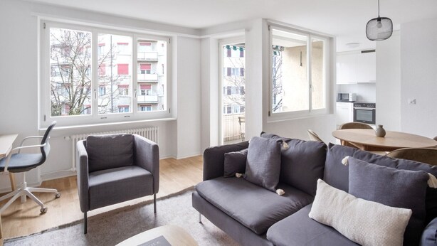Un salon moderne avec un canapé gris, une petite table et un fauteuil. De grandes fenêtres offrent une vue sur les bâtiments environnants.