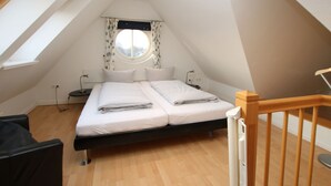 A small, single-room apartment with a sloped ceiling, a bed with white bedding, a round window with curtains, and a wooden floor. - Rungholtstraße 6, Whg. 4 - Apartment with 3 rooms on approx. 55m² for max. 2 persons. (Wyk auf Föhr)