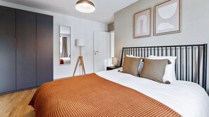 A modern bedroom with a large bed, a wardrobe, a bedside table, a floor lamp, and two framed pictures on the wall.