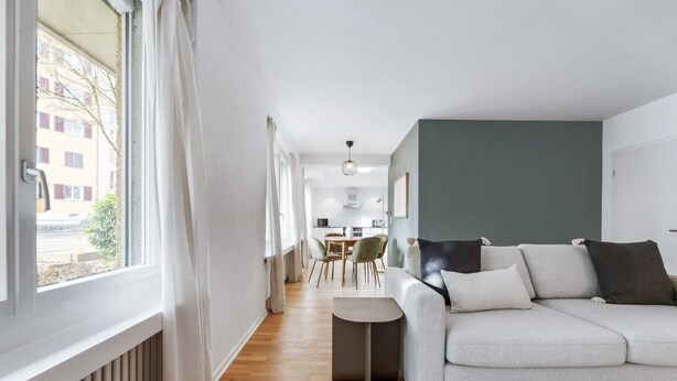 A modern living room with a sofa, a dining area, and a view of a building outside.
