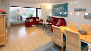 Un salon moderne avec un canapé rouge, une table à manger entourée de chaises en osier et une grande fenêtre donnant sur les bâtiments environnants.