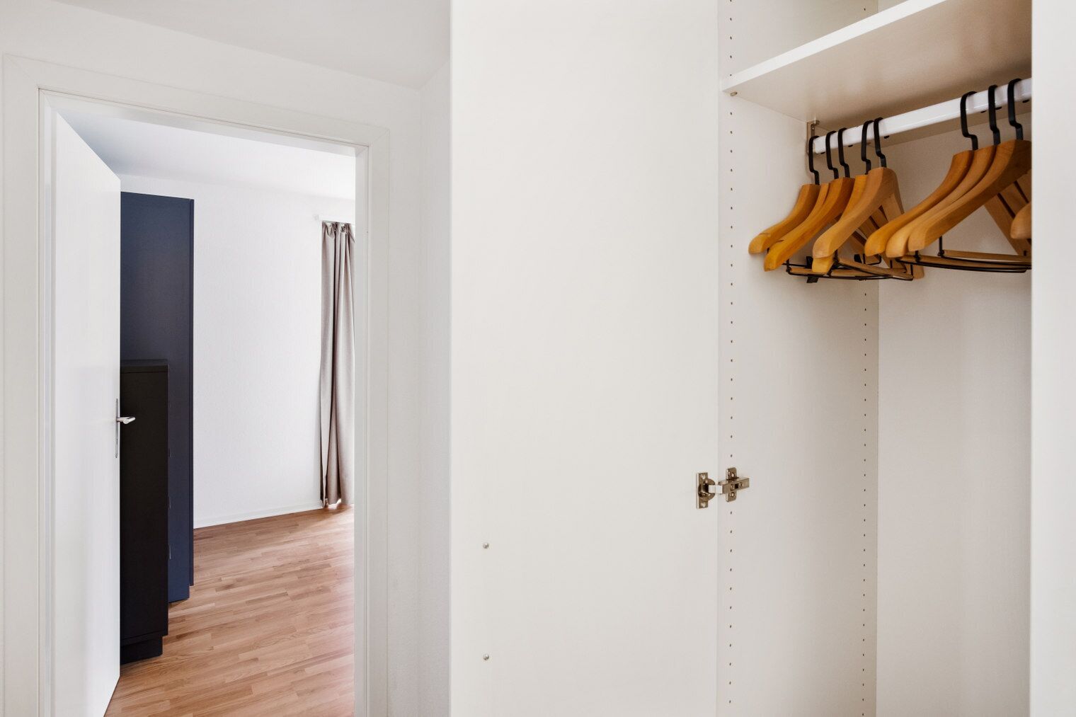 Un couloir doté d’un plancher en bois, d’une armoire blanche avec des cintres en bois, et d’une porte entrouverte donnant sur une autre pièce.