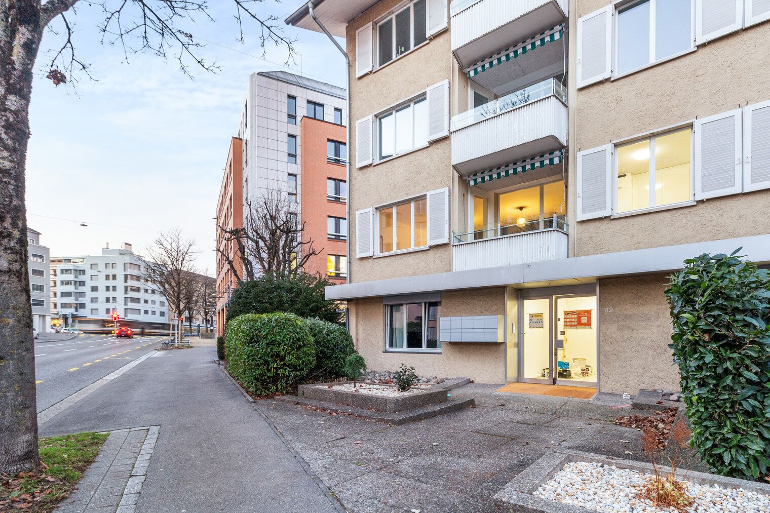 Un immeuble à plusieurs étages avec une entrée au niveau du sol, un petit espace jardin et un magasin visible au rez-de-chaussée.