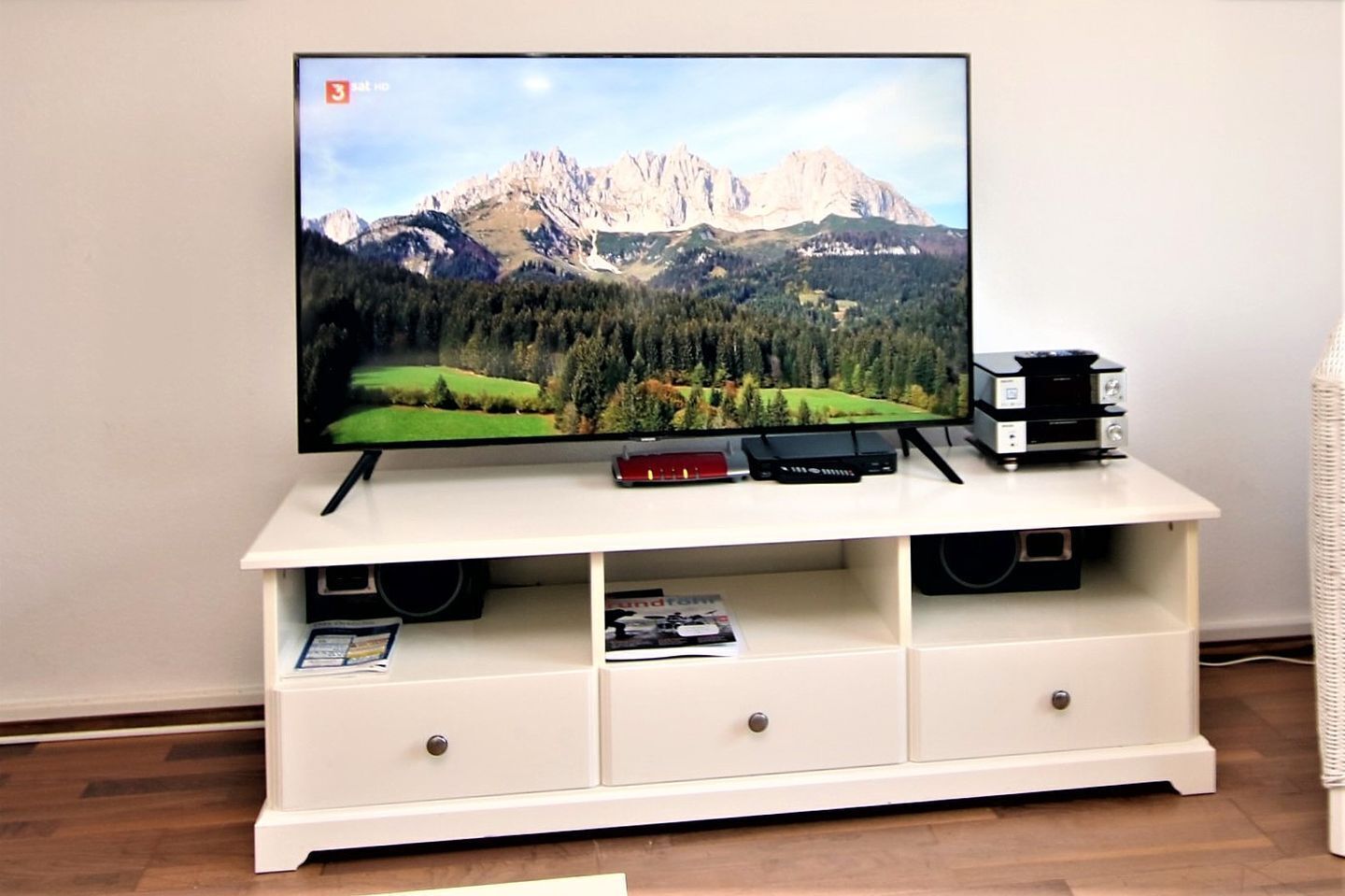 A flat-screen TV on a white entertainment center with two speakers, a remote, and a small book.