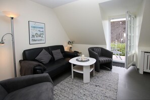 Una sala de estar moderna con un sofá de cuero negro, una mesa redonda blanca y un sillón gris. Un cuadro enmarcado cuelga de la pared, y una ventana ofrece vistas al exterior.