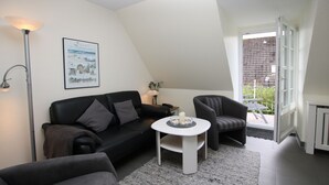 Ein modernes Wohnzimmer mit einer schwarzen Ledersofa, einem weißen runden Tisch und einem grauen Sessel. Ein gerahmtes Bild hängt an der Wand, und ein Fenster bietet einen Blick ins Freie.