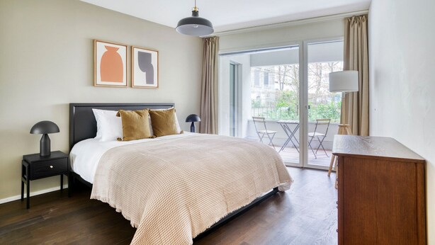 Een slaapkamer met een groot bed, een nachtkastje, een bureau, een stoel en een balkon met een tafel en stoelen.