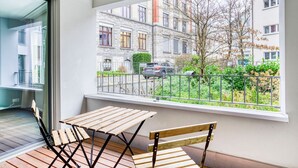 Een balkon met een houten tafel en stoelen, een metalen hekwerk en uitzicht op een woonwijk met gebouwen en een boom.