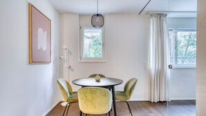 Een eetgedeelte met een ronde tafel en vier stoelen, een hanglamp, een lijst met een afbeelding aan de muur, en een raam met gordijnen.