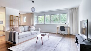 Een moderne woonkamer met een bank, salontafel en een bureau met stoel. Grote ramen zorgen voor natuurlijk licht en uitzicht op groen.