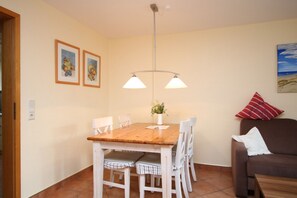 Een eetruimte met een houten tafel, witte stoelen, een hanglamp en een bruine bank met een rood kussen.