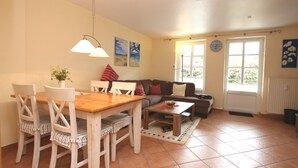 Een woonkamer met een eettafel, stoelen, een bank en een salontafel. Er zijn twee ramen met gordijnen, een radiator en een klok aan de muur.