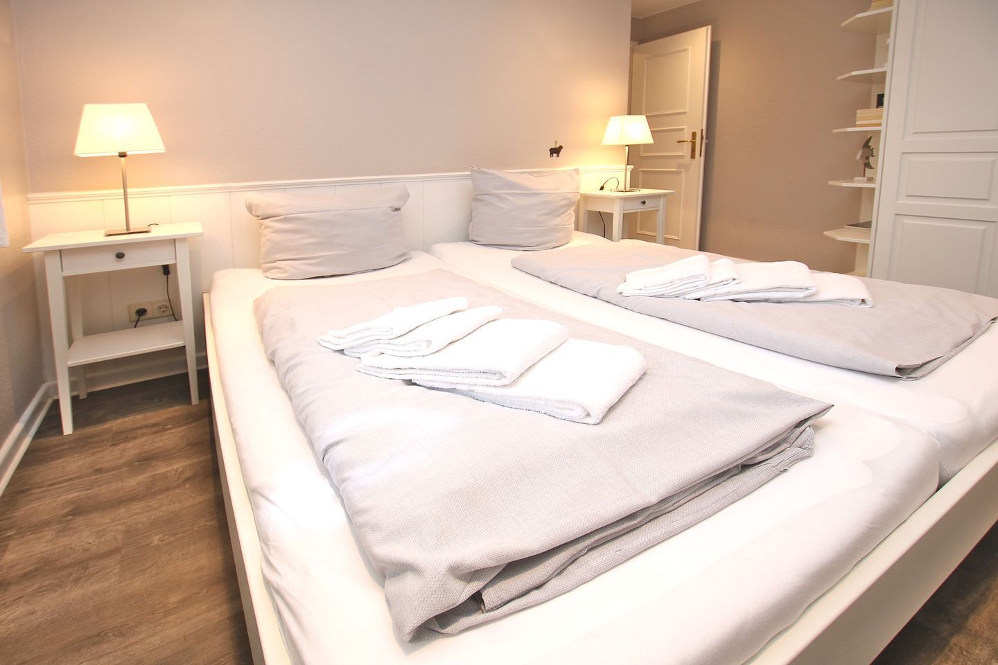 Ein Hotelzimmer mit zwei Betten, jeweils mit Kissen und gefaltetem Handtuch. An den Nachttischen stehen Lampen und eine kleine Ablagefläche.