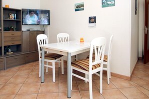Uma sala de jantar com uma mesa e cadeiras brancas, uma televisão na parede e uma estante com livros e objetos decorativos.