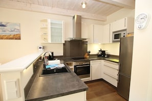 Een moderne keuken met een spoelbak, fornuis, oven en koelkast. Op de muur hangt een klok en boven de spoelbak een schilderij.