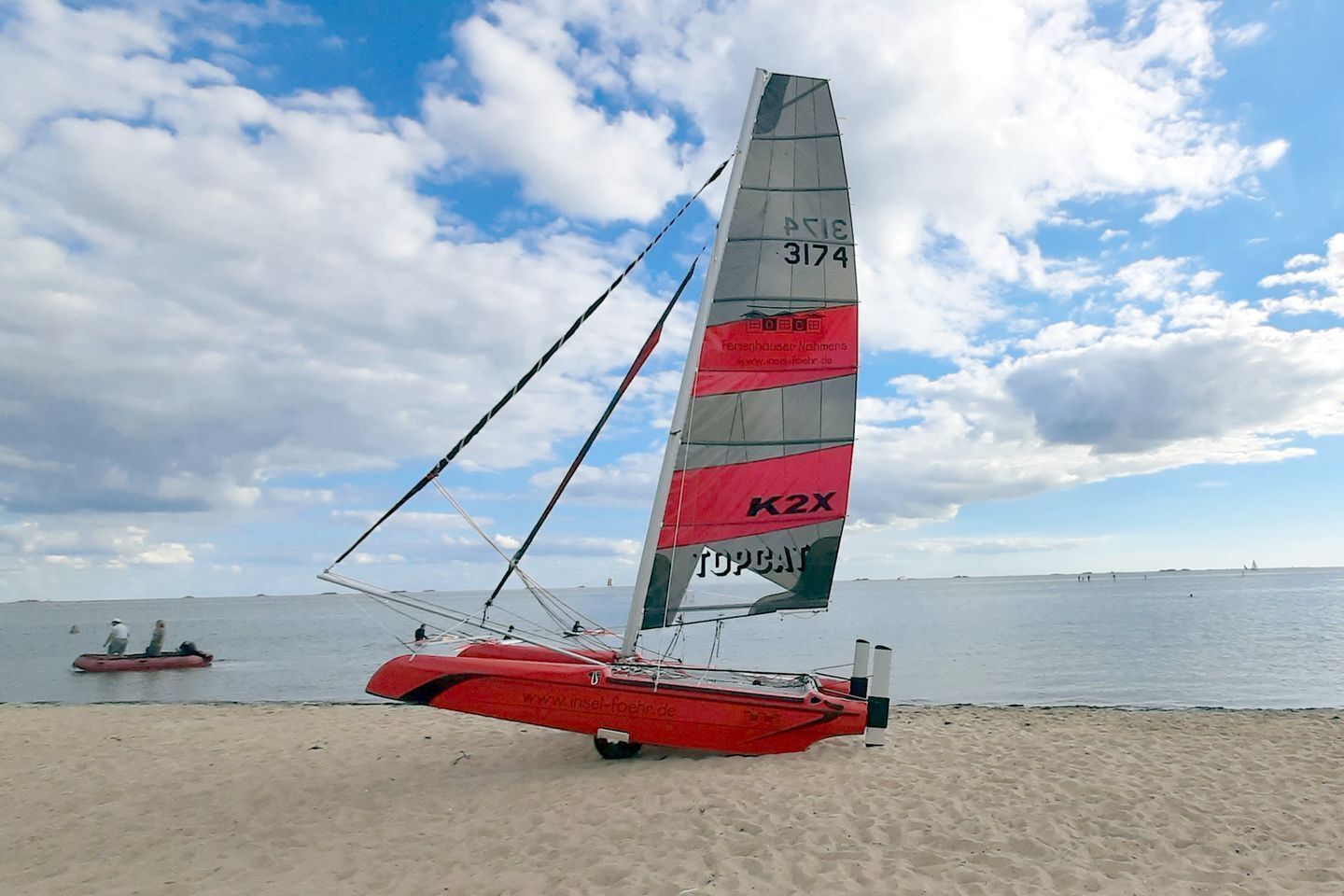 Un voilier rouge à la grande voile, sur une plage de sable.