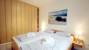 Een tweepersoonsbed met witte lakens, twee bedlampjes, een houten kledingkast en een schilderij van een strandlandschap aan de muur.