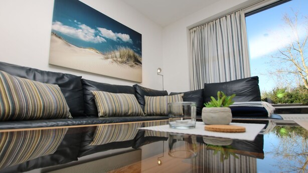Een moderne woonkamer met een leren zwarte bank, een glazen salontafel en een groot raam met gordijnen.