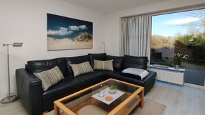 Een moderne woonkamer met een zwarte leren bank, een glazen salontafel, een groot raam met gordijnen en een schilderij aan de muur.