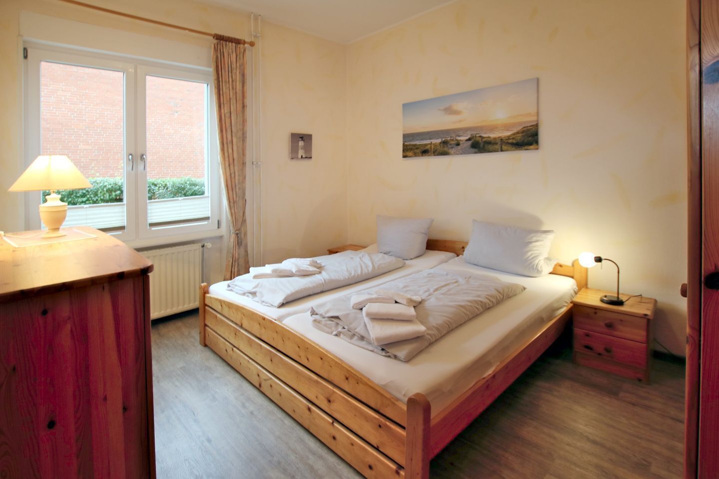 Ein Doppelbett mit weißer Bettwäsche, ein hölzerner Kopfteil, ein Nachttisch mit Lampe, ein Fenster mit Vorhängen und ein Gemälde an der Wand.