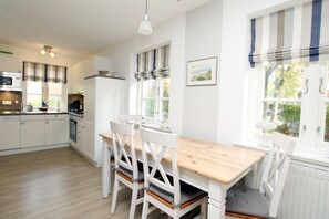 A modern kitchen with white cabinetry, a wooden dining table, and a window with striped curtains. - Strandstraße 8, Apt. 14, Green Eilun - Corner townhouse with approximately 75 m² spread over 4 floors (Wyk auf Föhr)