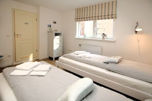 A hotel room with two single beds, a wardrobe, a radiator, a window with a striped valance, and a wall-mounted lamp. - Strandstraße 8, Apt. 14, Green Eilun - Corner townhouse with approximately 75 m² spread over 4 floors (Wyk auf Föhr)