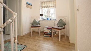 A hallway with two wooden chairs with cushions, a stack of books, a window with a striped valance, and a rope barrier. - Strandstraße 8, Whg. 14, Green Eilun - terraced end house with approx. 75m² over 4 floors (Wyk auf Föhr)
