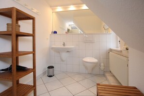 Une petite salle de bain avec un lavabo, des toilettes et un radiateur, tous de couleur blanche. On y trouve une étagère en bois avec des paniers et une poubelle.