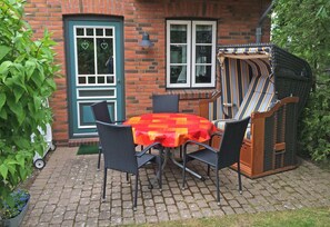 Une terrasse extérieure avec un mur en briques, une table avec des chaises et un fauteuil en osier doté d’un coussin à rayures.