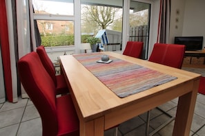 Un coin repas avec une table en bois, des chaises rouges, un set de table coloré et un téléviseur en arrière-plan.