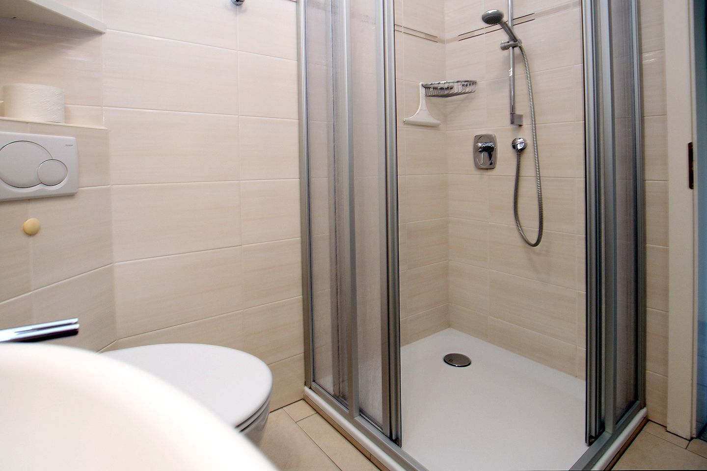 A glass-enclosed shower with a handheld showerhead, a wall-mounted faucet, and a small shelf.