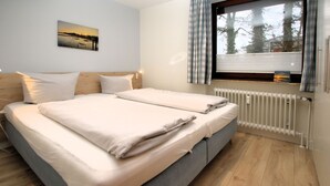 A double bed with a headboard, a window with curtains, a radiator, and a wall-mounted picture. - Badestraße 80, Whg. 1, Goting Hus - Newly renovated apartment on the mezzanine floor, (Wyk auf Föhr)