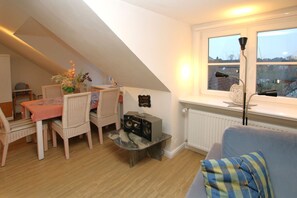 A cozy loft bedroom with a sloped ceiling, a dining area with wicker chairs, a table with a pink tablecloth, a radio, and a window with a view of buildings. - Mittelstraße 18, Whg. 1, Haus am Rosenbeet - Holiday apartment with 3 rooms on (Wyk auf Föhr)