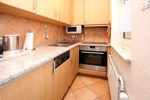 Una cocina compacta con armarios de madera, un lavamanos de acero inoxidable, microondas integrado, lavavajillas y horno.