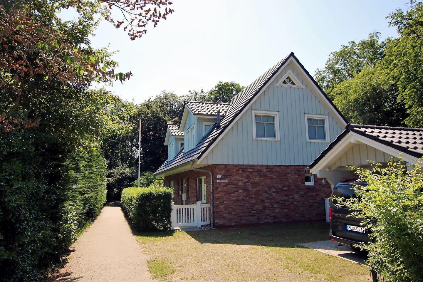 A two-story house with a brick foundation and a white upper section, a front porch, and a car parked in the driveway.