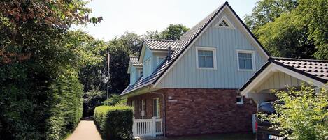 Une maison à deux étages, avec une fondation en briques et une partie supérieure blanche, un porche à l’avant et une voiture garée dans l’allée.