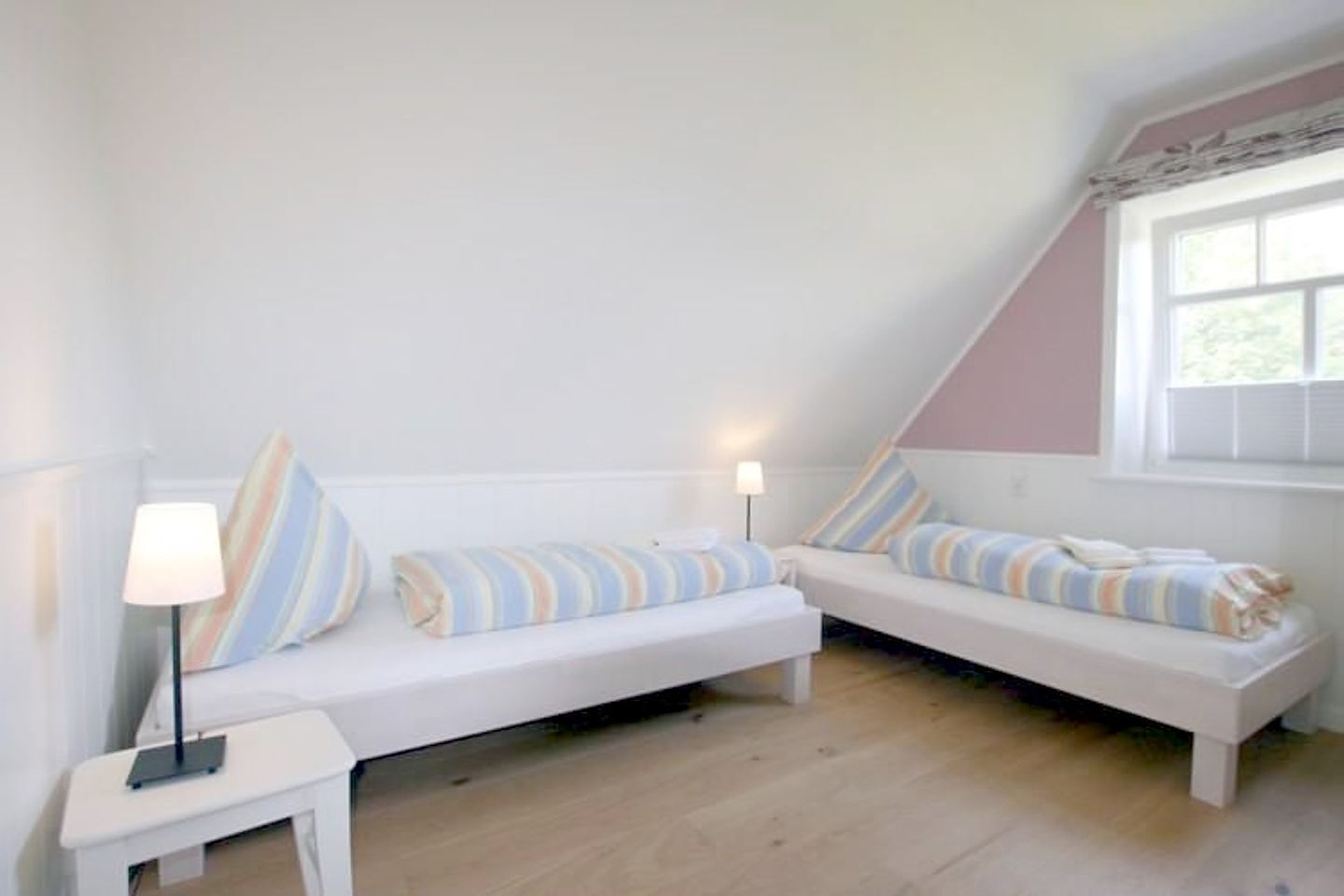 Een kamer met twee witte banken, gestreepte kussens, een tafeltje met een lamp en een raam met zonwering.