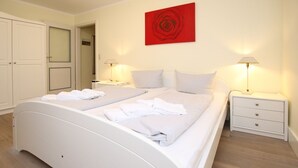 Un lit double avec du linge de lit blanc, deux tables de chevet avec des lampes, un tableau abstrait rouge accroché au mur et une porte avec un panneau en verre dépoli.